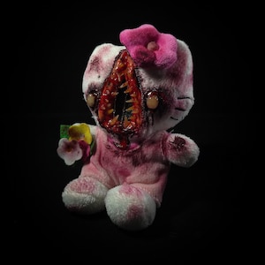 May include: A plush toy featuring a white and pink cat character with a large, vertical, tooth-filled mouth opening in the center of its face. It has a pink flower on its head, dark stained patches, and holds a small bouquet of colorful fabric flowers.