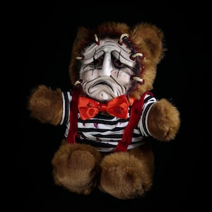 May include: A brown teddy bear with a distressed face, stitched around the edges. It is dressed in a black and white striped top, red braces, and a red bow tie. The bear has brown fur and is set against a black backdrop.