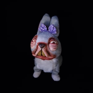 May include: A plush, light blue rabbit with a purple bow. The rabbit has exaggerated features, including large, red-rimmed eyes and a wide, open mouth with visible teeth. The toy is set against a black background.