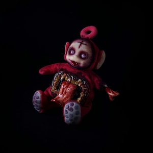 May include: A red plush toy with a disturbing, horror-themed design. The toy has a cross on its forehead, a gaping wound exposing internal organs, and purple eyes. The background is solid black.