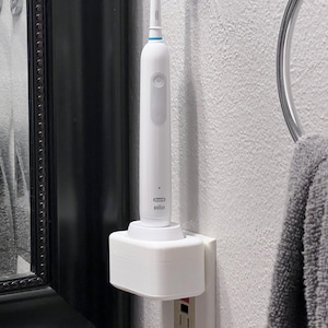 May include: A white electric toothbrush with a green and white brush head is docked in a white charging base. The base is plugged into a wall outlet. The toothbrush has a power button and the brand name "Oral-B".