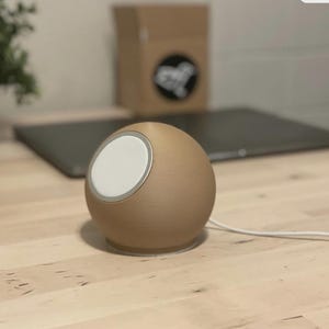 May include: A spherical, tan-colored charging station with a white circular charging pad and a silver trim. A white charging cable extends from the base. The device sits on a wooden surface, with a laptop and a box in the background.