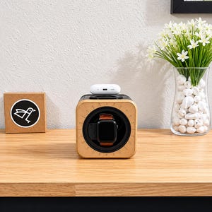 May include: A wooden watch winder with a black smartwatch and brown leather band. A white wireless charging case sits on top. A small cardboard box with a bird logo is to the left. A vase of white flowers is to the right.