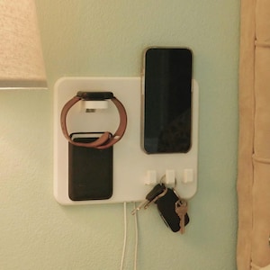 May include: A white wall-mounted organizer holds a smartphone, smartwatch, and keys. The smartwatch has a brown band. The smartphone and a small rectangular item are black. Keys hang below, and a charging cable is visible.