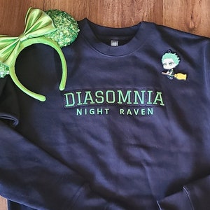 Può includere: Felpa nera con testo ricamato in verde che recita "Diasomnia Night Raven". Un personaggio dei cartoni animati verde con una scopa è ricamato sulla parte superiore destra della felpa.