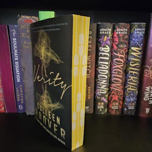 May include: A yellow and black paperback book titled "Verity" by Colleen Hoover. The book is on a bookshelf with other books, including "Rebel Witch" and "Belladonna" by Kristen