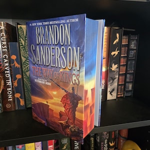 May include: A hardcover book titled "The Way of Kings" by Brandon Sanderson is displayed on a black bookshelf. The book has a blue and orange cover with a warrior illustration. Other books are visible on the shelf.