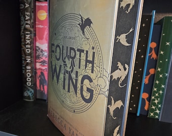 May include: A gold and black book with the title "Fourth Wing" and the text "Fly...or Die" on the cover. The book is on a bookshelf with other books.