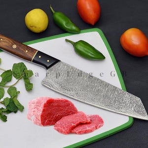 May include: A Damascus steel chef's knife with a wooden handle and black bolster rests on a cutting board with sliced raw meat, tomatoes, chillies, and a lemon. The knife has a patterned blade, designed for kitchen use.