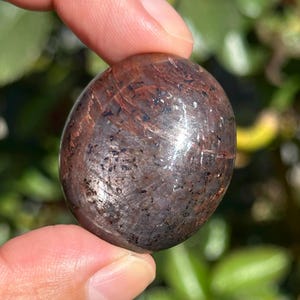 May include: A polished, oval-shaped gemstone with a deep reddish-brown base color, featuring dark, scattered inclusions and a smooth, reflective surface that catches the light, displaying subtle internal textures and natural mineral patterns.