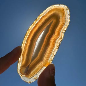 May include: A translucent, oval-shaped agate slice with bands of orange, brown, and white. The natural stone is held against a bright blue sky, showcasing its layered patterns and unique texture. The agate is a decorative item.