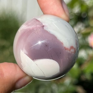 May include: A polished sphere featuring a blend of colors. The orb showcases a mix of white and lavender hues, with subtle cracks and variations in the stone. The sphere is held between fingers, with a blurred background.