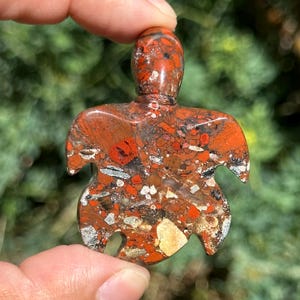 May include: Red, brown, and white speckled stone turtle pendant. The turtle is carved in a stylized shape with a flat back and a rounded front.