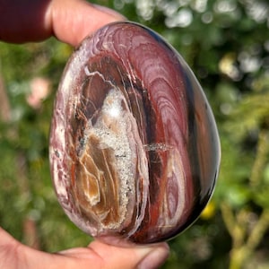 May include: A polished, egg-shaped stone with a swirling pattern of red, brown, white, and black hues. The stone's surface is smooth and reflective, showcasing the natural variations in color and texture. The stone is held in a hand.
