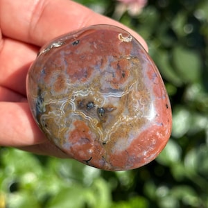 May include: A polished, oval-shaped stone with a mix of colors. The stone features a base of reddish-brown with patterns of tan, white, and dark blue. The surface is smooth and reflective, with natural variations in color and texture.