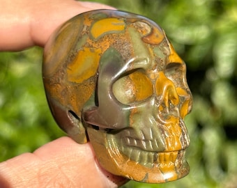 May include: A carved skull made of green, brown, and yellow jasper. The skull has a detailed face with a prominent nose and teeth.