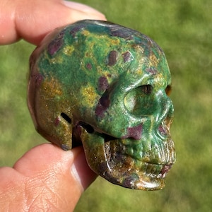 May include: A green and brown skull carved from a stone with red and gold flecks.