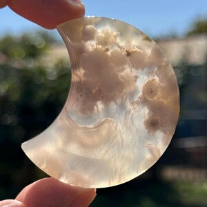 May include: A crescent moon-shaped gemstone with a translucent, milky appearance. The stone features swirling patterns of beige and cream, with a polished surface. The gemstone is held up against a blurred outdoor background.