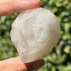 May include: A clear quartz crystal carved into the shape of an alien skull. The skull has detailed features, including eyes, a nose, and a mouth.