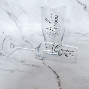 May include: Two clear glass drinking glasses with black lettering. One glass says "John Groom" and the other says "Alex Bride" with a heart symbol.