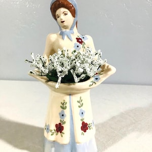 May include: A ceramic figurine of a person wearing a light blue bonnet and a cream-coloured coat with red and blue floral patterns. The figure holds a wide, shallow tray filled with a cluster of small white artificial flowers in their hands.