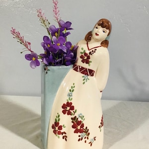 Puede incluir: Un jarrón de cerámica con forma de mujer con un vestido blanco con diseños florales rojos y azules. El jarrón tiene una base rectangular azul y blanca. La mujer sostiene un ramo de flores moradas.