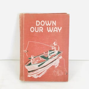 Puede incluir: Libro vintage titulado "Down Our Way" con una portada roja. La portada presenta una ilustración blanca de dos personas pescando desde un barco. El título del libro está en letras blancas.