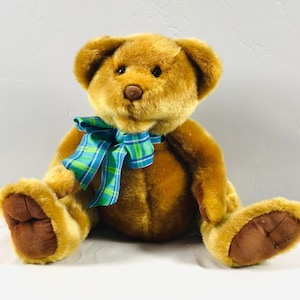 May include: A brown teddy bear with a blue and green plaid bow tie. The bear is sitting on a white surface.