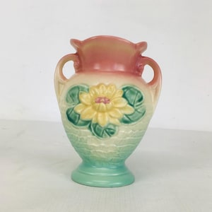 Puede incluir: Un jarrón de cerámica vintage con un diseño de dos tonos. La parte superior es de color rosa apagado, que pasa a un color crema con una flor amarilla y hojas verdes. La base es de un verde suave y el jarrón tiene dos asas.