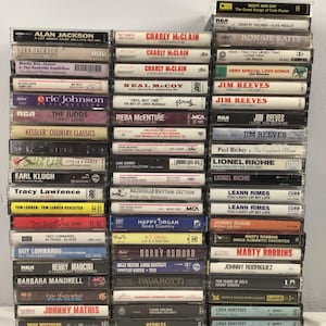 Könnte beinhalten: Ein Stapel alter Musikkassetten mit verschiedenen Künstlern. Die Kassetten haben Etiketten mit Künstlernamen und Albumtiteln, darunter Alan Jackson und Jim Reeves. Die Hüllen sind aus transparentem Kunststoff.