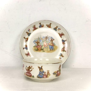 Puede incluir: Un juego de cuenco y plato de cerámica vintage con un tema de conejitos. El cuenco y el plato blancos tienen ribetes dorados. El plato tiene una imagen central de conejitos y un borde de conejitos. El cuenco tiene un patrón repetido de conejitos.