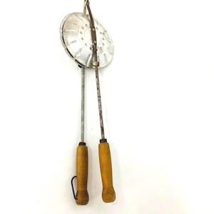 Op de afbeelding: Vintage metalen taartijzer met houten handvatten. Het taartijzer heeft een cirkelvormig kookoppervlak met een scharnierend ontwerp. De handvatten zijn gemaakt van lichtgekleurd hout en vertonen tekenen van slijtage. Een kleine metalen lus is aan één handvat bevestigd.