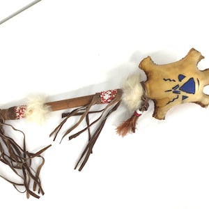 Puede incluir: Un sonajero ceremonial nativo americano con una cabeza de cuero crudo y un mango de madera. La cabeza es de color canela con diseños geométricos azules. El mango está envuelto con cuentas rojas y blancas, piel blanca y flecos de cuero.