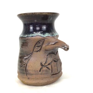 May include: A handmade brown ceramic pitcher with a spout and handle. The pitcher features carved leaf and geometric designs. The rim is dark brown, transitioning to a deep blue and light green glaze. The pitcher is a unique piece of pottery.