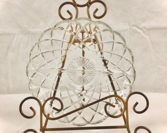 May include: A clear glass serving dish with a diamond pattern and a floral center, displayed on a decorative bronze metal stand. The stand has scrolled details and a curved design, adding an elegant touch to the presentation.