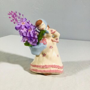 Puede incluir: Figura de cerámica de una mujer sosteniendo un ramo de flores moradas. La figura viste un vestido color crema con detalles y ribetes florales rosas. El sombrero de la mujer y la cesta que sostiene las flores son azules.