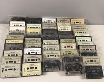 May include: A collection of vintage audio cassette tapes in clear plastic cases. The tapes are arranged in rows and feature various titles and artists.