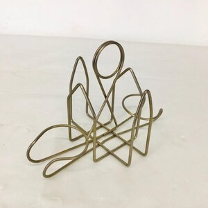 May include: A vintage brass wire napkin holder. The holder features a geometric design with a circular handle and angular shapes. The brass has a warm, metallic tone, and the design is open and airy.