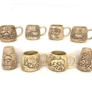 May include: A collection of eight beige ceramic mugs and a vase, each with a unique embossed design. The mugs feature scenes of cabins, landscapes, and maps. The vase has a floral design. All items have a rustic, vintage aesthetic.