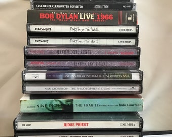 May include: A vertical stack of seventeen music CDs in plastic jewel cases. The spines display titles including Aerosmith, Bad Company, Eric Clapton, Creedence Clearwater, Bob Dylan, Pink Floyd, Jerry Garcia, Van Morrison, Nine Inch Nails, Judas Priest, Led Zeppelin, Lynyrd Skynyrd, and Billy Squier.