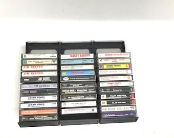 May include: A black plastic storage rack holding three vertical columns of music cassette tapes. The collection includes various artists such as Jim Reeves, Marty Robbins, Randy Travis, Leann Rimes, Clay Walker, and Yanni, with visible text on each spine.
