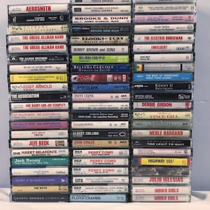 May include: A stack of vintage cassette tapes featuring various artists, including Aerosmith, Brooks & Dunn, Belinda Carlisle, and more. The tapes are arranged vertically, with the titles and artist names visible on each cassette.