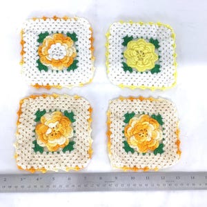 May include: Four handmade crochet coasters. Each coaster features a square design with a white background, a central flower motif in shades of yellow and orange, and green leaves. The edges are trimmed with yellow or orange.