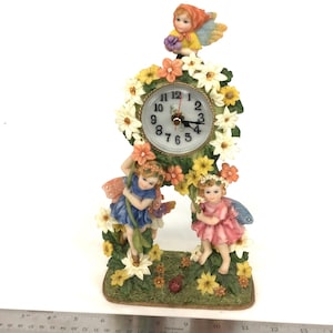 May include: A decorative clock featuring a floral design and three fairy figurines. The clock face is surrounded by white and yellow flowers. The fairies are dressed in blue and pink, with wings. The clock is approximately 15 cm tall.