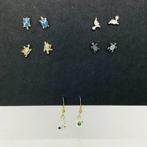 May include: A collection of five pairs of turtle-shaped earrings. The set includes two pairs of gold-tone stud earrings with blue and gold enamel, one pair of silver-tone studs, one pair of black studs, and one pair of clear glass drop earrings.
