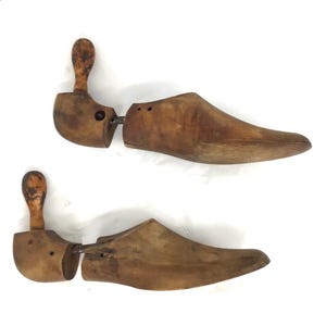 May include: Two antique wooden shoe stretchers. Each stretcher has a wooden handle, a curved heel piece, and a shoe-shaped front. The wood has a warm, aged tone, suggesting vintage craftsmanship. The stretchers are designed to maintain the shape of shoes.