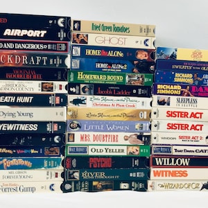 Op de afbeelding: Een stapel VHS-banden met filmtitels als "Airport", "Home Alone" en "Sister Act". De banden zijn in verschillende kleuren, met titels gedrukt in vette, contrasterende lettertypen. De collectie bevat familiefilms en actiefilms.