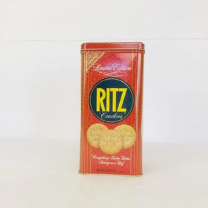 Puede incluir: Una lata rectangular roja y dorada de galletas Ritz. La lata presenta el logotipo de Ritz en amarillo y azul, con una imagen de tres galletas. El texto en la lata dice "Edición Limitada" y "Todo sabe mejor sentado en un Ritz."