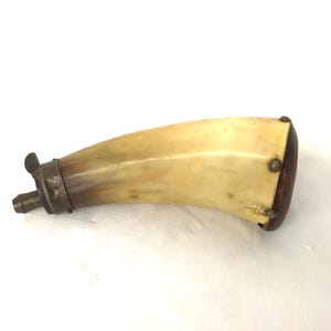 May include: A powder horn, likely for black powder, with a curved, off-white body and a dark brown wooden base. The horn has a metal spout and metal bands around the top. The horn is likely an antique.