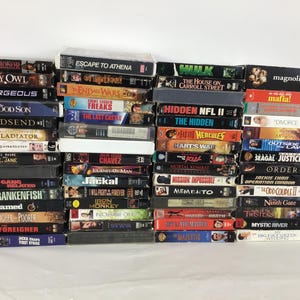 Puede incluir: Una colección de cintas VHS, con títulos como "Guns of Honor", "Escape to Athena" y "Magnolia". Las cintas están apiladas, mostrando una variedad de géneros cinematográficos y portadas coloridas. Sobre una superficie blanca.
